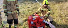 Zdjęcie przedstawia trzech strażaków podczas ćwiczeń. Jeden ze strażaków obsługuje motopompę.