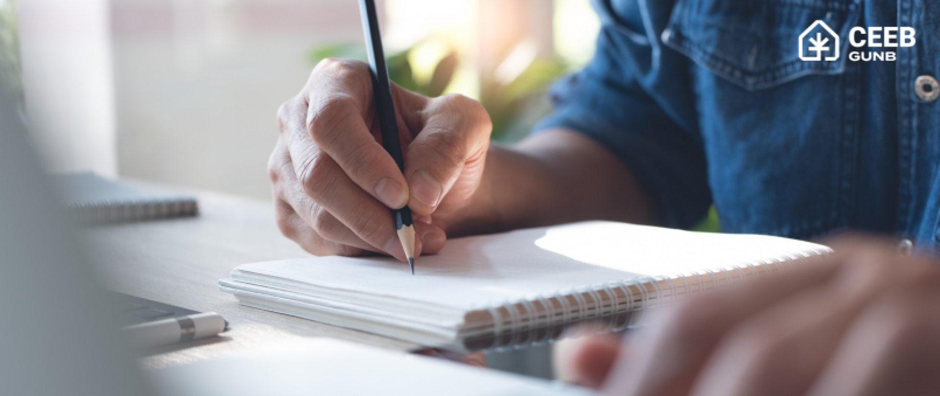 Person writing in a notebook with a pencil, wearing a blue denim shirt; another hand visible in the foreground. Indoor setting with natural light and a "CEEB GUNB" logo in the top right corner.