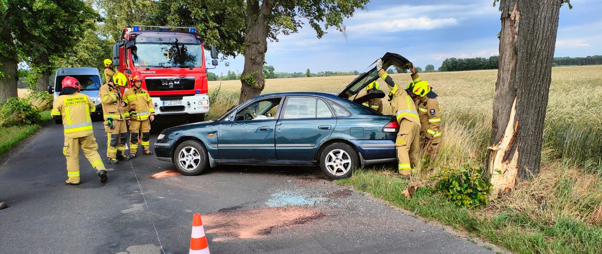 Samochód osobowy stoi na jezdni obok pracują strażacy w tel samochody strażackie.