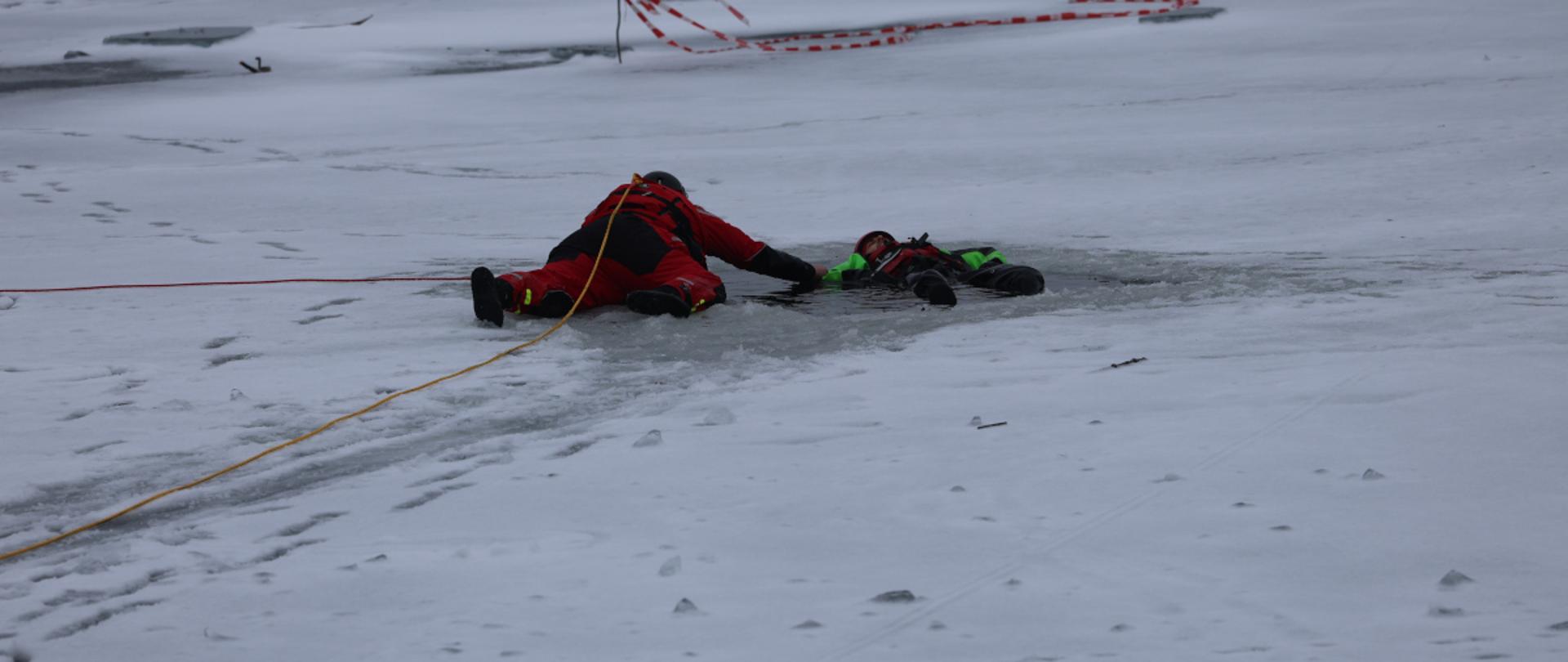 Dwóch ratowników w specjalistycznych kombinezonach leży na lodzie przy przeręblu. Jeden z nich wyciąga drugiego z wody, trzymając go za rękę, a do ratowników przymocowana jest lina asekuracyjna. W tle widoczna jest taśma ostrzegawcza i zamarznięta powierzchnia zbiornika.