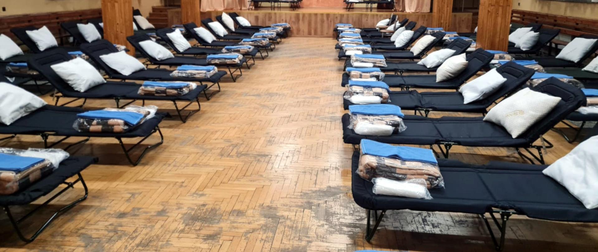 Na zdjęciu widoczna jest sala w budynku ochotniczej straży pożarnej. Po obydwu stronach w czterech rzędach ustawione są łóżka polowe ,na nich leżą zapakowane koce, ręczniki i poduszki.