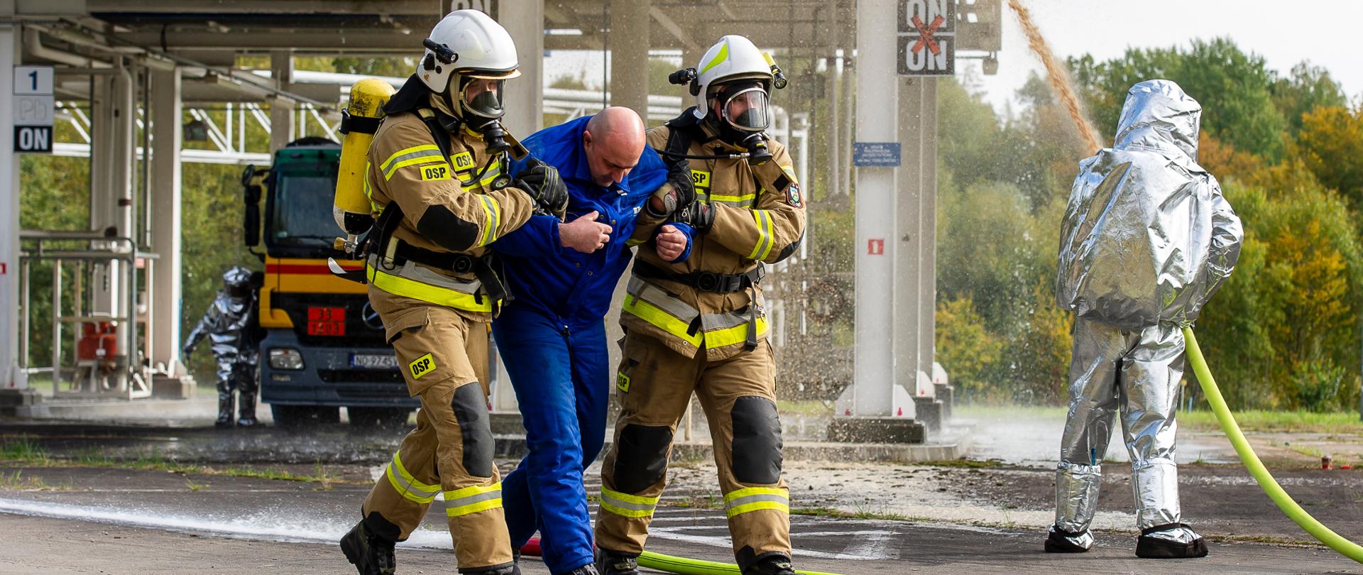 Rota strażaków ewakuuje osobę poszkodowaną, Z lewej strony strażak w ubraniu żaroochronnym przeszukuje miejsce wypadku. Z prawej strony widoczna sylwetka strażaka podającego w natarciu prąd ciężkiej piany gaśniczej na cysternę z paliwem.