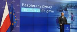 Minister Grzegorz Puda przemawia podczas konferencji, w tle ścianka z napisem "Bezpieczny pieszy 100 mln zł dla gmin".