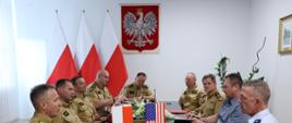 Gabinet komendanta głównego. Spotkanie kierownictwa komendy głównej z przedstawicielkami amerykańskiej gwardii narodowej. Na zdjęciu na pierwszym planie widoczny jest stół konferencyjny przy którym siedzą: komendant główny z zastępcą, dyrektorzy biur oraz dwuosobowa amerykańska delegacja. Na stole widoczne są proporce z flagami. W tle na ścianie widoczne godło i trzy polskie flagi. 