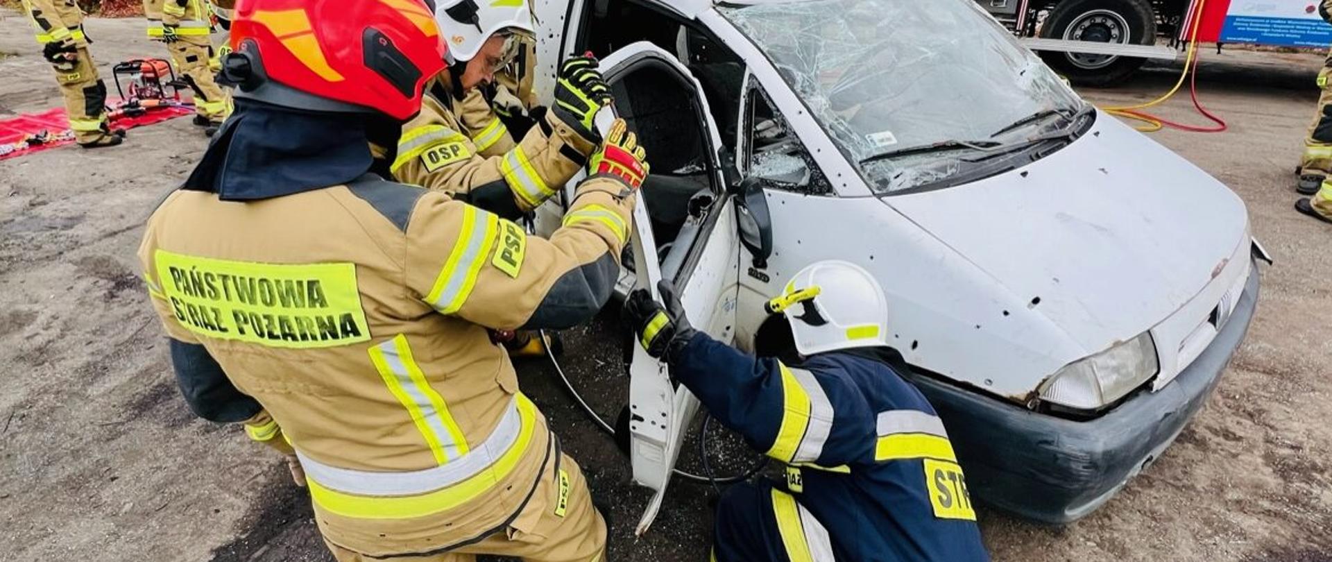 Na zdjęciu strażacy w umundurowaniu bojowym otwierający drzwi białego samochodu dostawczego. W tle czerwony samochód strażacki.