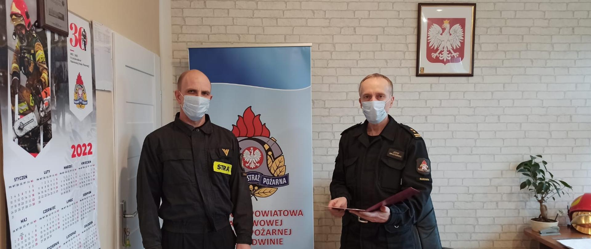 Zdjęcie przedstawia dwóch mężczyzn ubranych w czarne mundury służbowe oraz maseczki chirurgiczne, jeden z nich trzyma w rękach dokumenty. Za nimi widnieje baner z logiem PSP oraz napisem: "Komenda Powiatowa Państwowej Straży Pożarnej w Świdwinie". Za banerem widzimy ścianę z tapetą w szare cegły. Na ścianie wisi Godło Rzeczypospolitej Polskiej. Zdjęcie wykonano w gabinecie Komendanta Powiatowego PSP w Świdwinie.