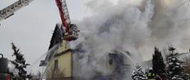 Widoczny pożar budynku mieszkalnego, silne zadymienie, podnośnik hydrauliczny i strażacy pracujący na nim