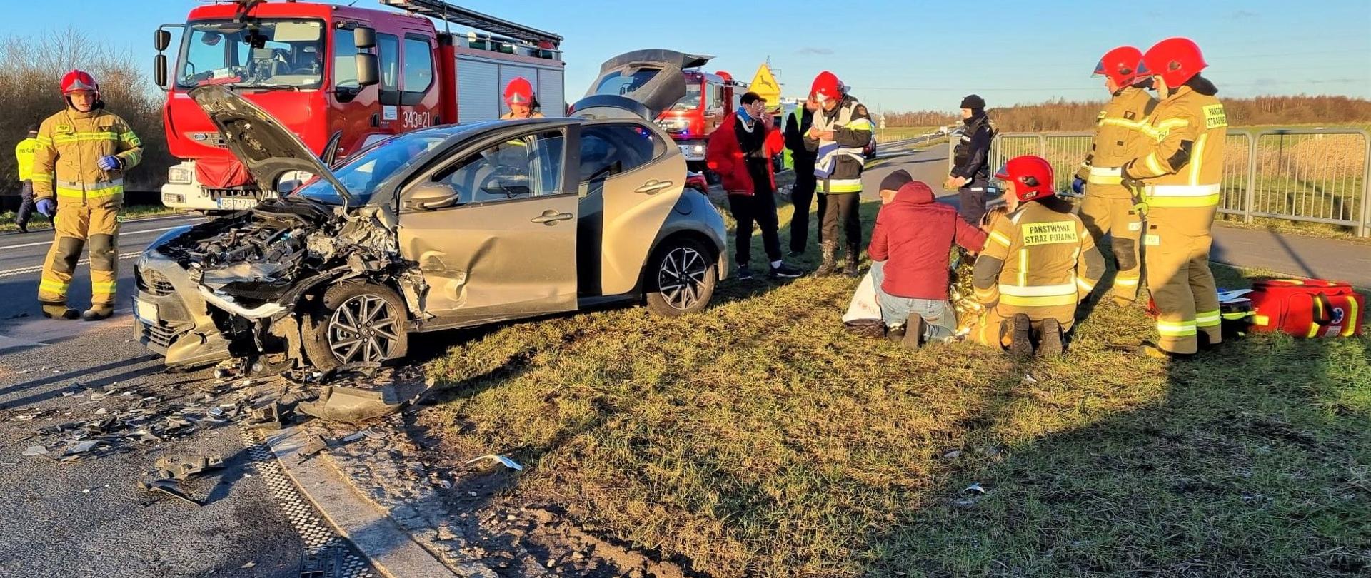 Zdjęcie przedstawia strażaków udzielających pomocy podczas wypadku. Na zdjęciu także uszkodzone auto.