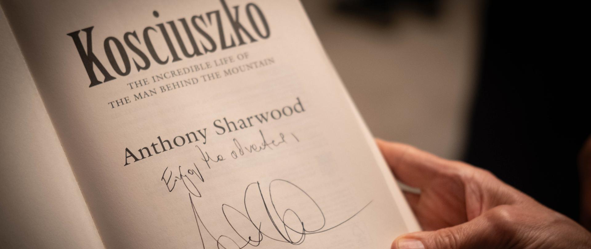 A book held in hands, open on the first page with the title and the author's autograph