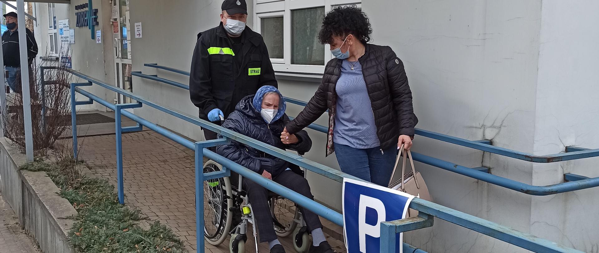 Na zjeździe z punktu szczepień, strażak w ubraniu koszarowym prowadzi na wózku inwalidzkim starszą osobę. Obok idzie kobieta, która trzyma za rękę osobę na wózku. 