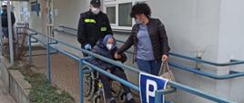 Na zjeździe z punktu szczepień, strażak w ubraniu koszarowym prowadzi na wózku inwalidzkim starszą osobę. Obok idzie kobieta, która trzyma za rękę osobę na wózku. 