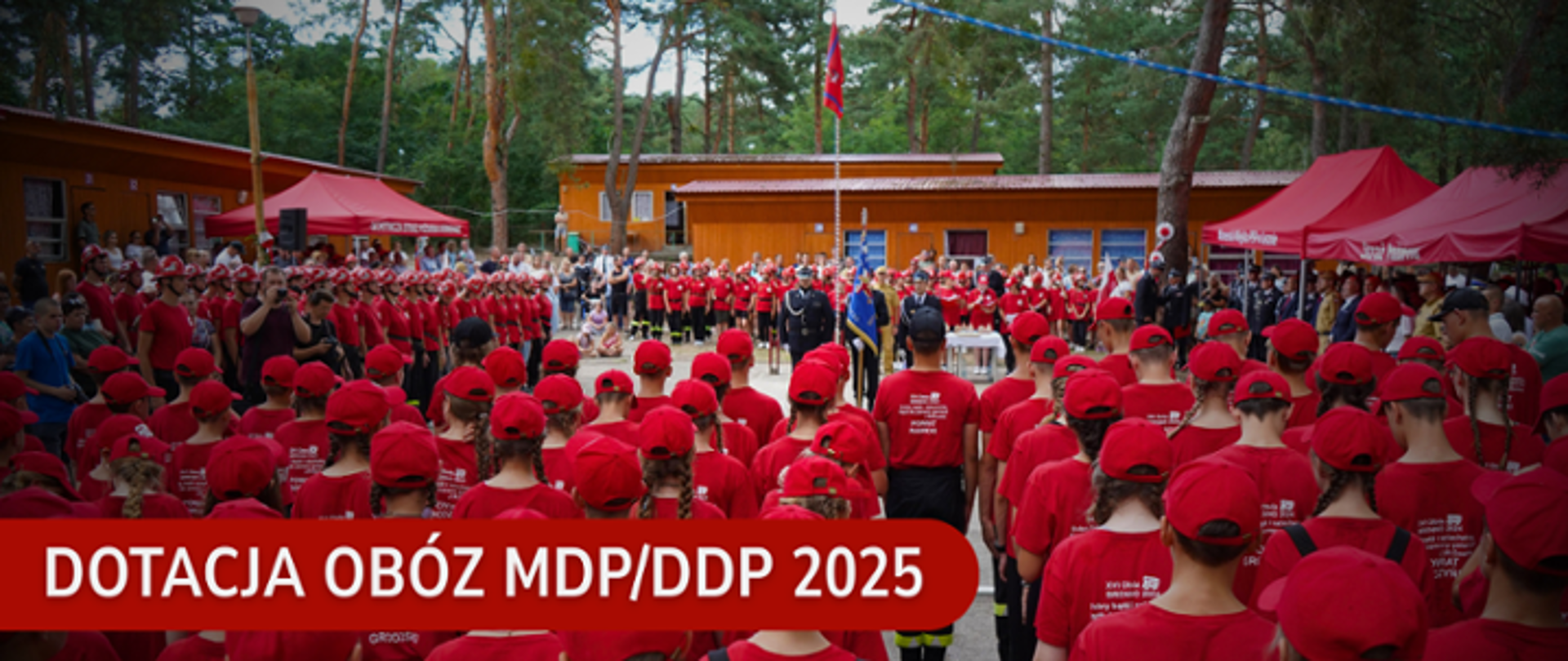Zbiórka młodzieżowych drużyn pożarniczych (MDP/DDP) na obozie w 2025 roku. Uczestnicy ubrani w czerwone koszulki i czapki stoją na leśnym placu przed drewnianymi budynkami. W tle widoczne czerwone namioty i maszt z flagą. Na grafice znajduje się napis: „Dotacja Obóz MDP/DDP 2025”.