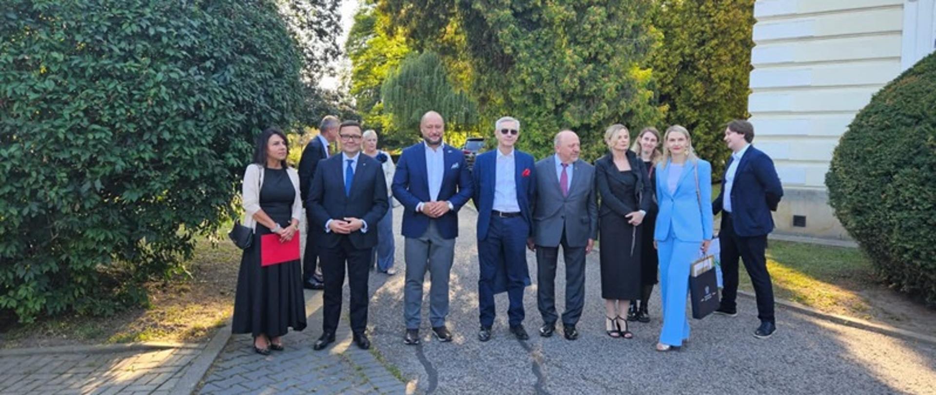 W parku przy Pałacu Radziwiłłów stoją w rzędzie elegancko ubrani przedstawiciele instytucji samorządowych, rolniczych itp. Od lewej strony stoi kobieta o ciemnych włosach w czarnej długiej sukience i jasnej marynarce, która trzyma w rękach czerwoną teczkę. Po jej prawej stronie stoi 4 mężczyzn w jasnych koszulach, krawatach i ciemnych garniturach. Pierwszy z nich to dyrektor Instytutu Zootechniki PIB – Tomasz Jacek. Obok mężczyzn stoi kobieta o blond włosach, w czarnej długiej sukience i czarnym żakiecie, którą jest dyrektor Oddziału Regionalnego KRUS w Krakowie – Iwona Szablińska, a obok niej stoi kobieta o blond włosach, ubrana w jasny niebieski garnitur. W ich otoczeniu rosną drzewa, przez które przebijają się promienie słońca. Po prawej stronie widoczny jest fragment budynku Pałacu Radziwiłłów w kolorze jasnym żółtym. Osoby te stoją częściowo na asfaltowej drodze, a częściowo na kostce brukowej.