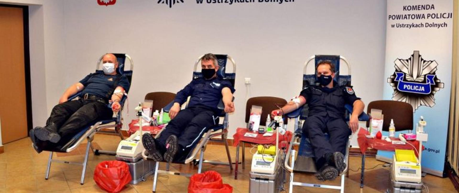 Na zdjęciu zastępca komendanta powiatowego psp w ustrzykach dolnych wraz z komendantami policji, oddają krew. w tle logo Komendy Powiatowej Policji 