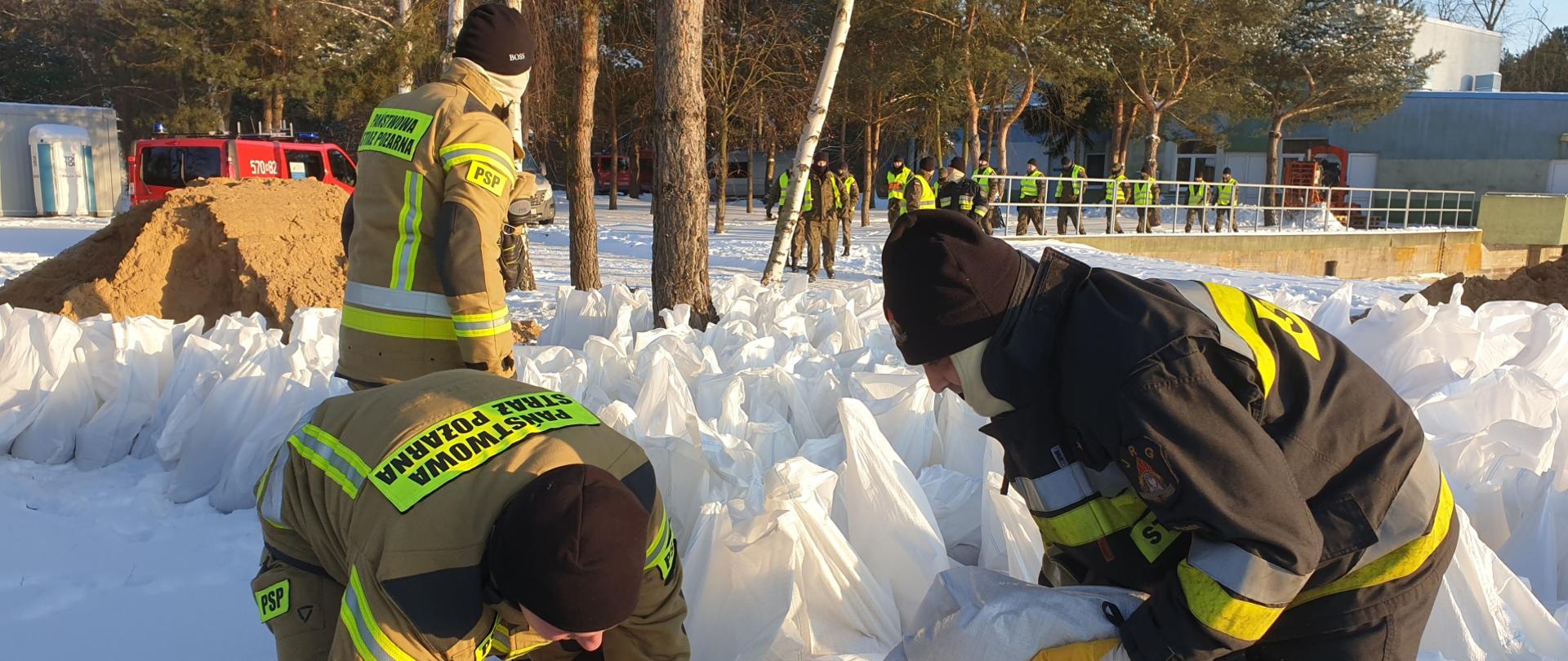 Strażacy w ubraniach specjalnych układający worki z piaskiem. w tle idą osoby ubrane w urania odlaskowe.
