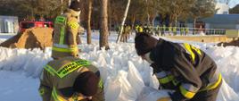 Strażacy w ubraniach specjalnych układający worki z piaskiem. w tle idą osoby ubrane w urania odlaskowe.