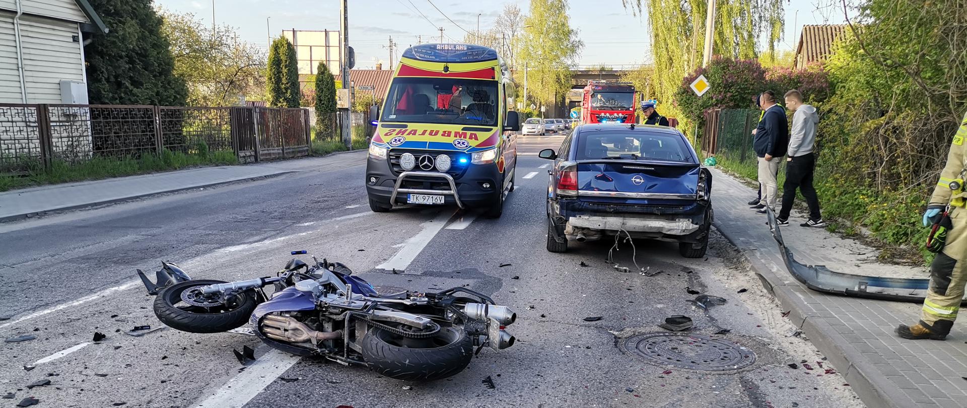 Zdjęcie przedstawia ulicę Malików. Na jezdni leży przewrócony, zniszczony motocykl, a przed nim samochód osobowy. Przy aucie, po lewej stronie stoi ambulans Ratownictwa Medycznego, a w głębi widać samochód pożarniczy.