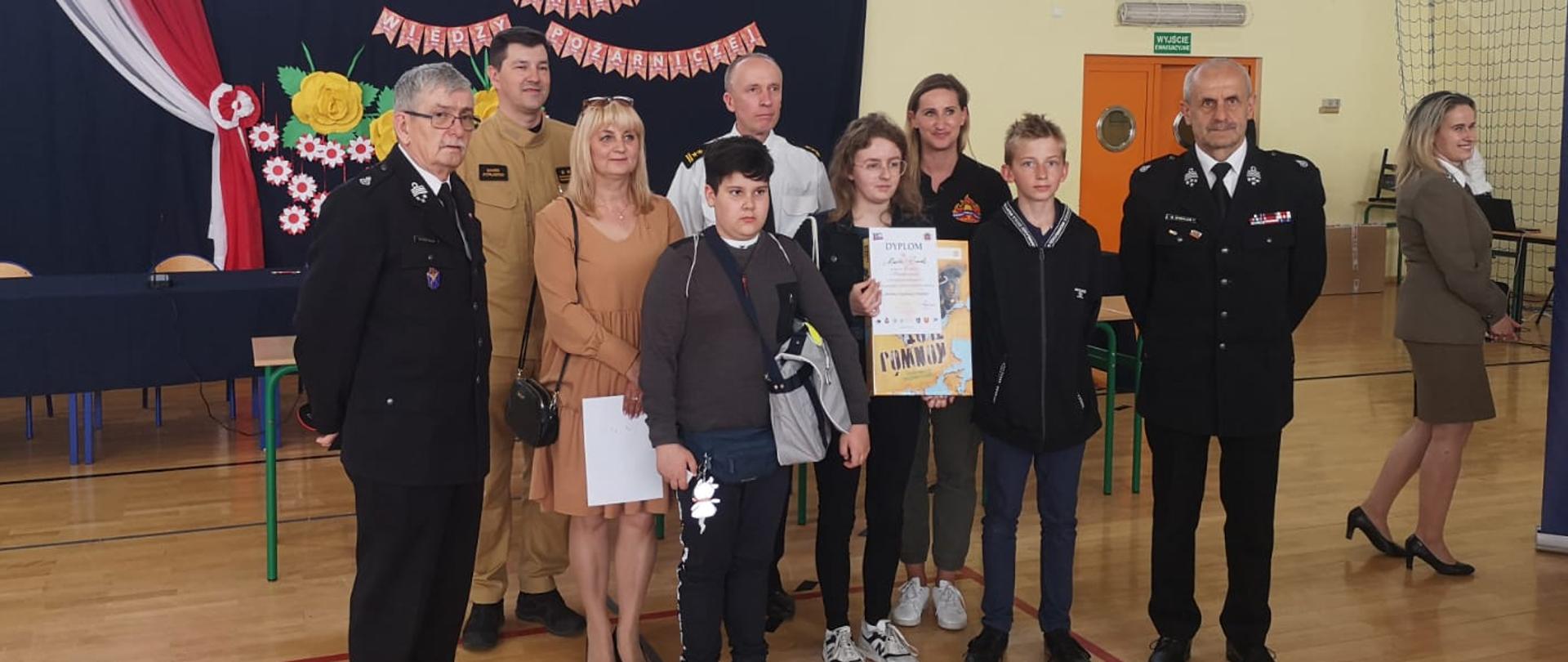 Zdjęcie przedstawia halę sportową na której rozgrywane są eliminacje wojewódzkie do Ogólnopolskiego Turnieju Wiedzy Pożarniczej pod hasłem "Młodzież zapobiega pożarom". Na hali pod banerem ustawieni są uczestnicy i organizatorzy turnieju pozujący do zdjęcia.