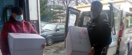 Na pierwszym planie dwaj przedstawiciele jednego z domów pomocy społecznej trzymają w rękach pudełka z maseczkami ochronnymi. Na drugim planie widoczny samochód dostawczy. 