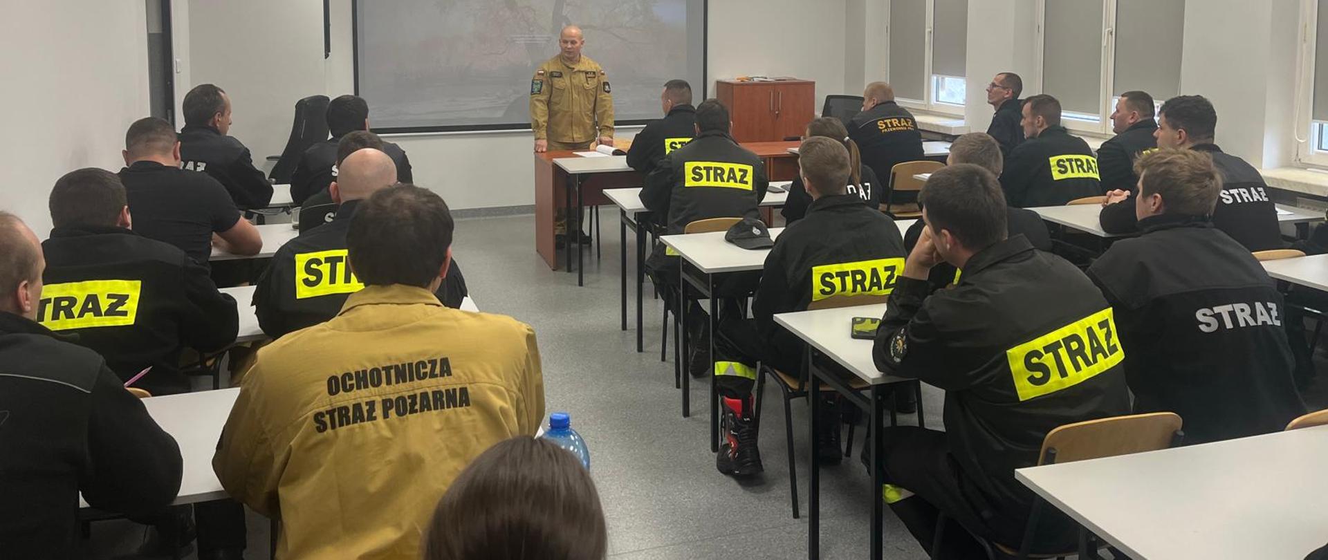 Instruktor z PSP prowadzi zajęcia teoretyczne dla uczestników szkolenia kierujących działaniem ratowniczym w sali wykładowej Ośrodka Szkolenia KW PSP w Opolu. Strażacy w mundurach słuchają prezentacji.