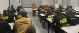 Instruktor z PSP prowadzi zajęcia teoretyczne dla uczestników szkolenia kierujących działaniem ratowniczym w sali wykładowej Ośrodka Szkolenia KW PSP w Opolu. Strażacy w mundurach słuchają prezentacji.
