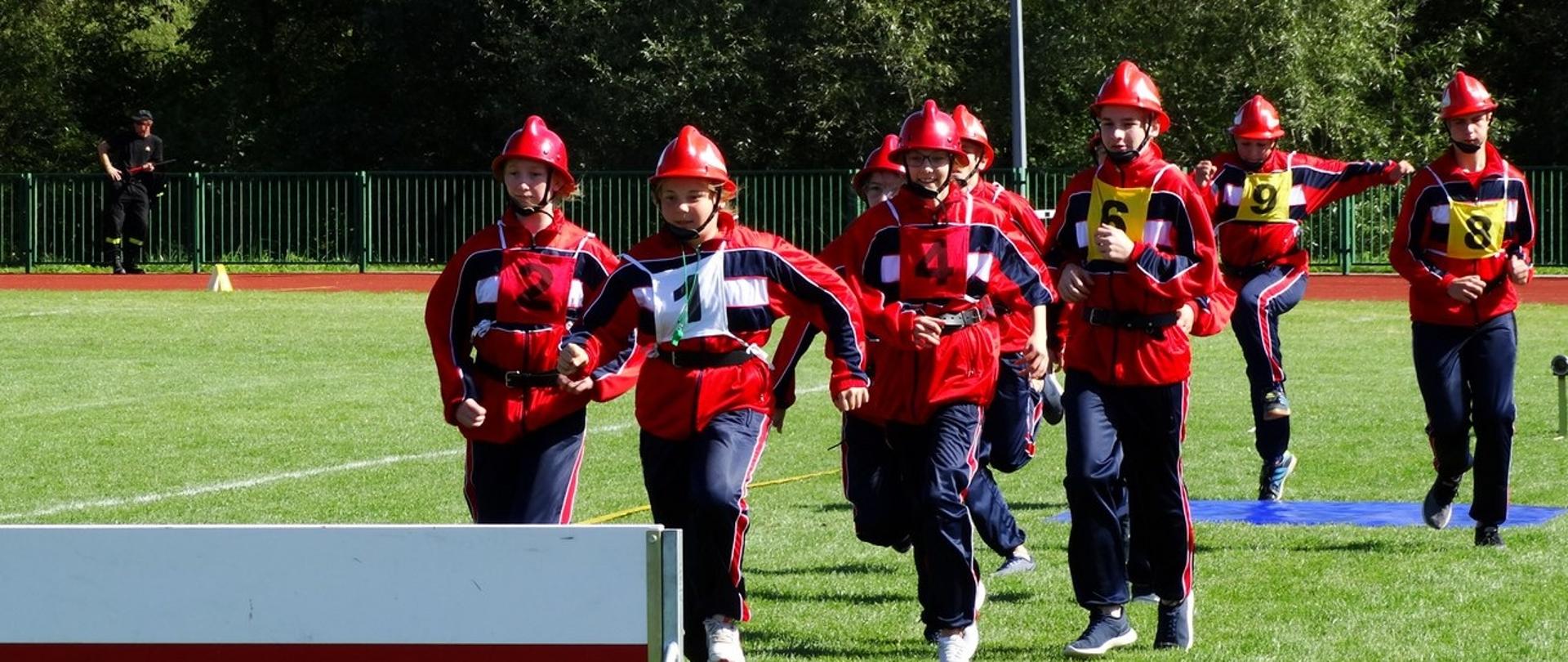 Na zdjęciu drużyna dziewcząt ubrana w czerwono-granatowe dresy biegnie w stronę przeszkody