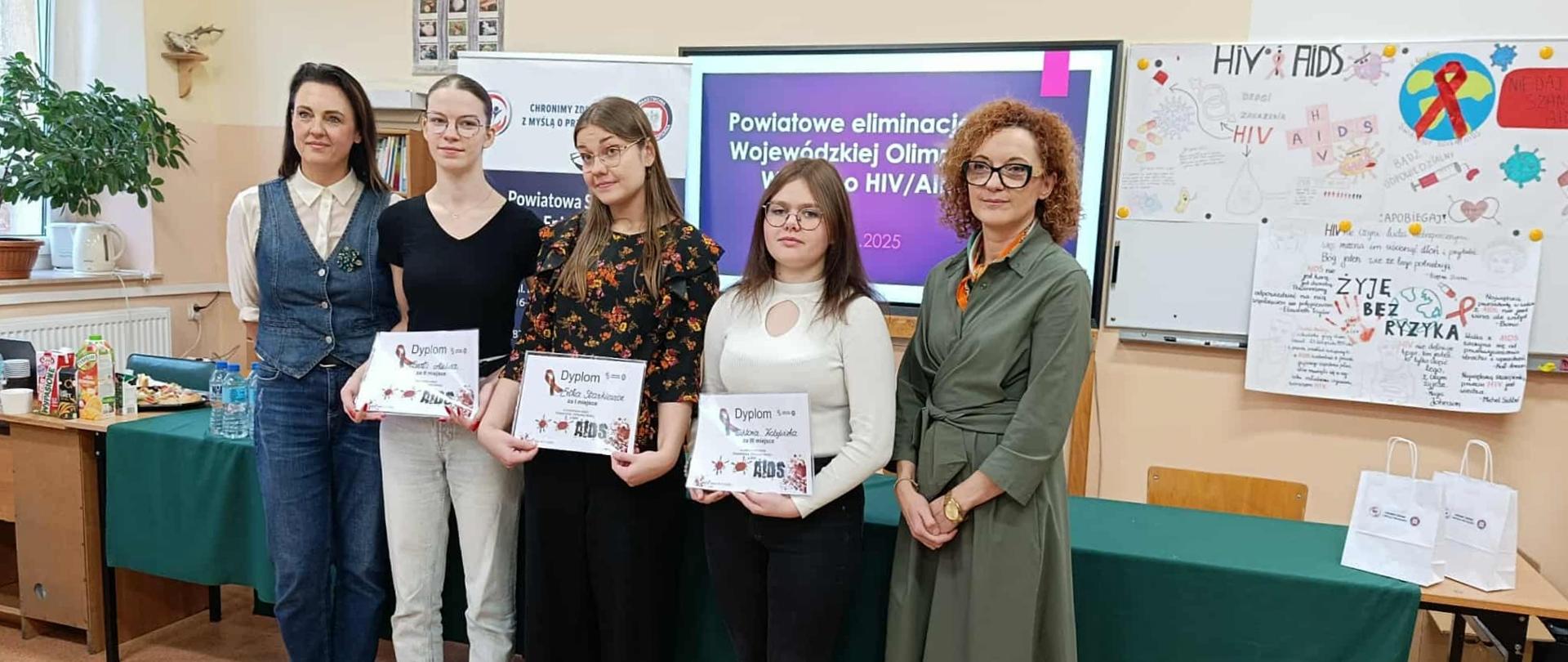 Pięć kobiet, w tym trzy uczennice trzymające dyplomy za udział w powiatowych eliminacjach Wojewódzkiej Olimpiady Wiedzy o HIV/AIDS, pozuje do zdjęcia w sali lekcyjnej. W tle widoczna jest prezentacja i tablica z informacjami na temat AIDS.
