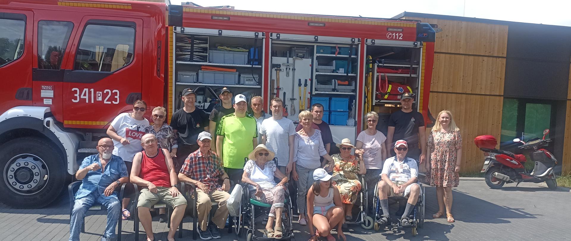 Grupa osób cywilnych wraz ze strażakami na tle wozu strażackiego.