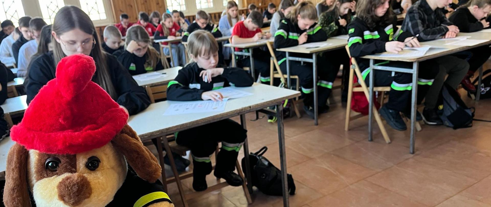 Na pierwszym planie maskotka strażak w oddali widać siedzące przy stolikach w trzech rzędach dzieci, które rozwiązują testy. 
