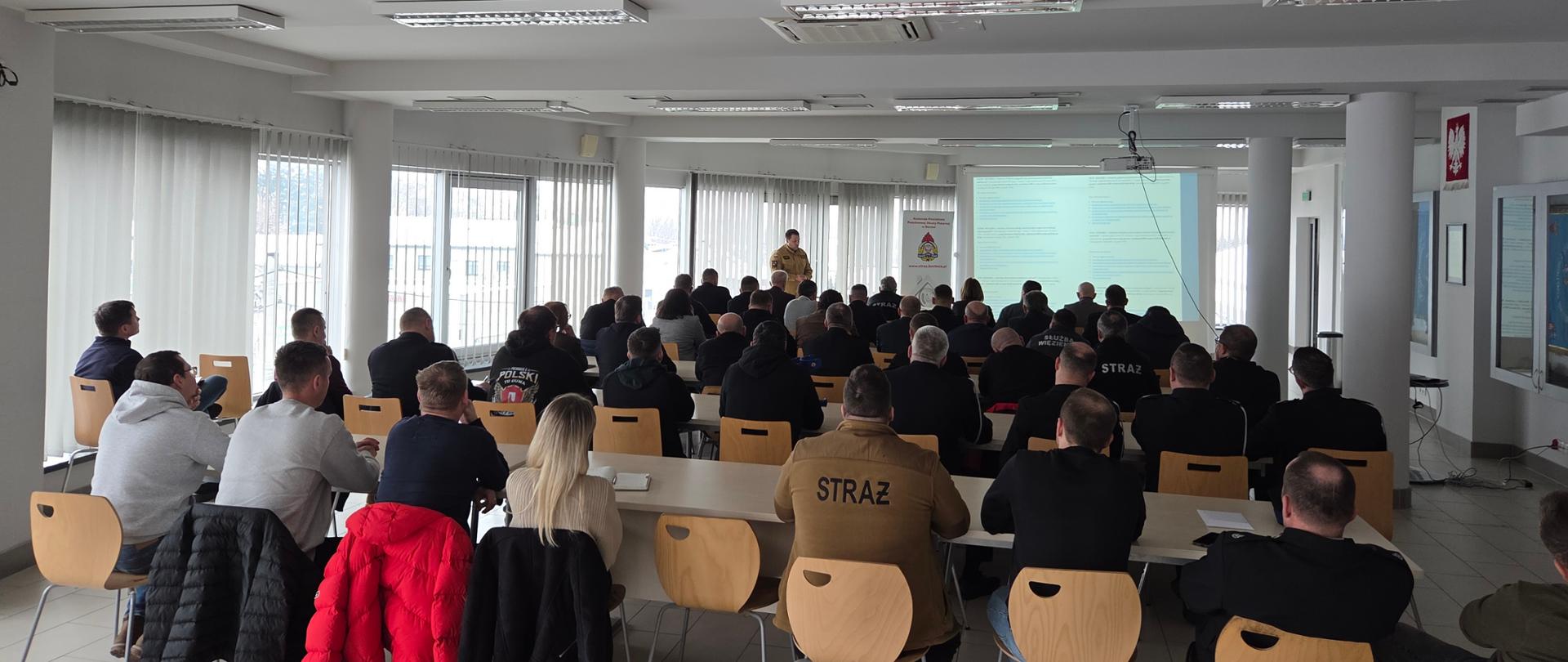 Grupa ludzi znajduje się na dużej sali. Przed grupą ludzi wyświetlana jest informacja na ekranie, jeden strażak stoi, pozostali ludzie siedzą na krzesłach w ławkach. Część ludzi posiada umundurowania różnych służb (straż, służba więzienna) pozostałe osoby w mundurach wyjściowych lub ubraniach cywilnych. 