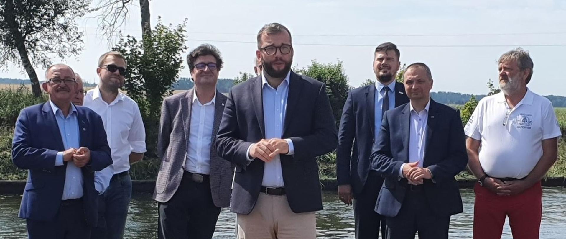 Na zdjęciu widać się osiem osób stojących na tle jeziora, wśród nich znajduje się minister Grzegorz Puda 