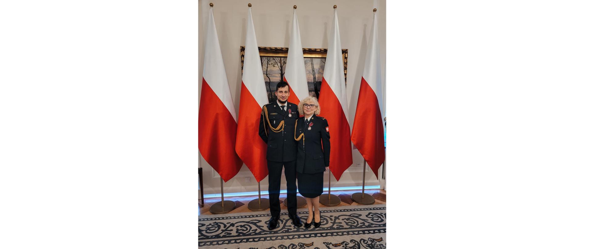 Strażacy w mundurach wyjściowych stoją pozują do zdjęcia. W tle biało czerwone flagi