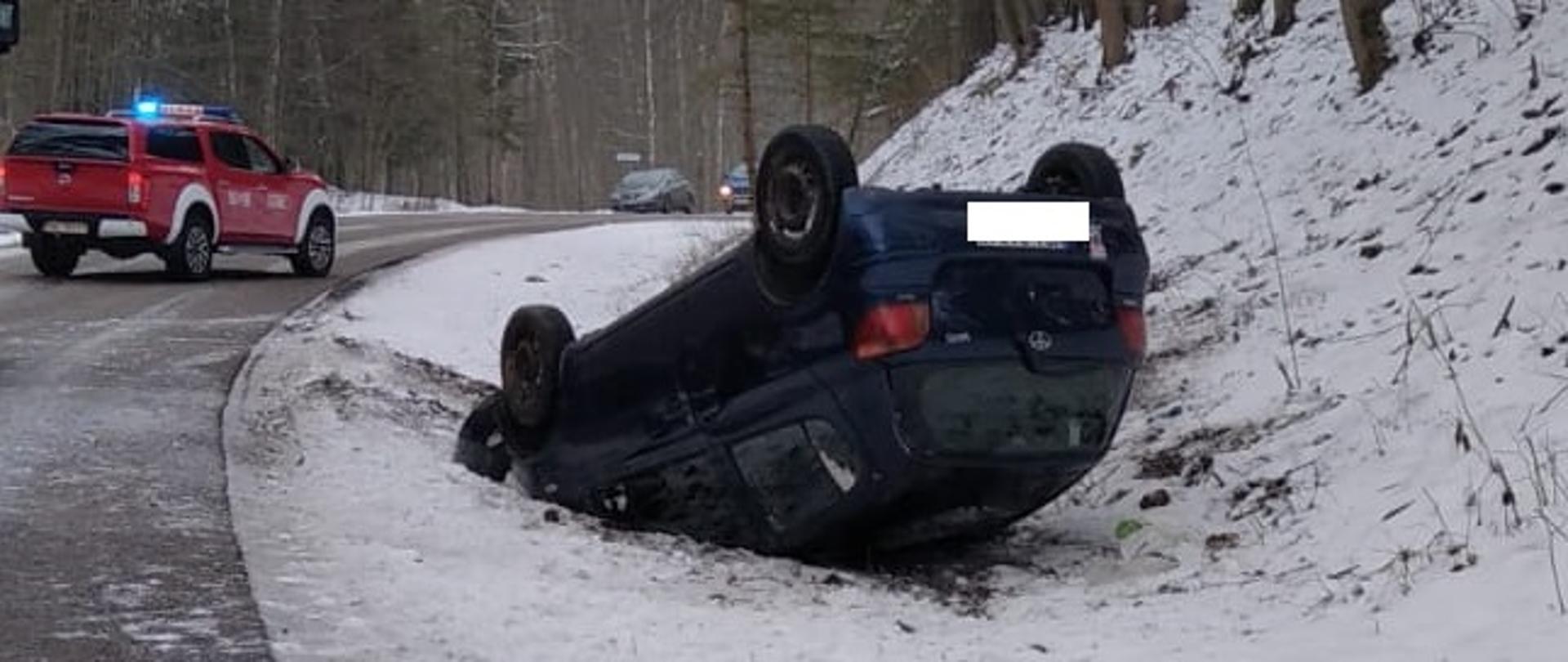 Samochód osobowy leży na dachu w rowie. Za nim w oddali wdać samochód PSP oraz inne samochody osobowe. 