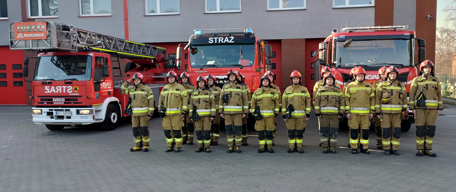 Grupa strażaków w umundurowaniu specjalnym stoi w dwuszeregu na tle samochodów pożarniczych z włączonymi światłami ostrzegawczymi