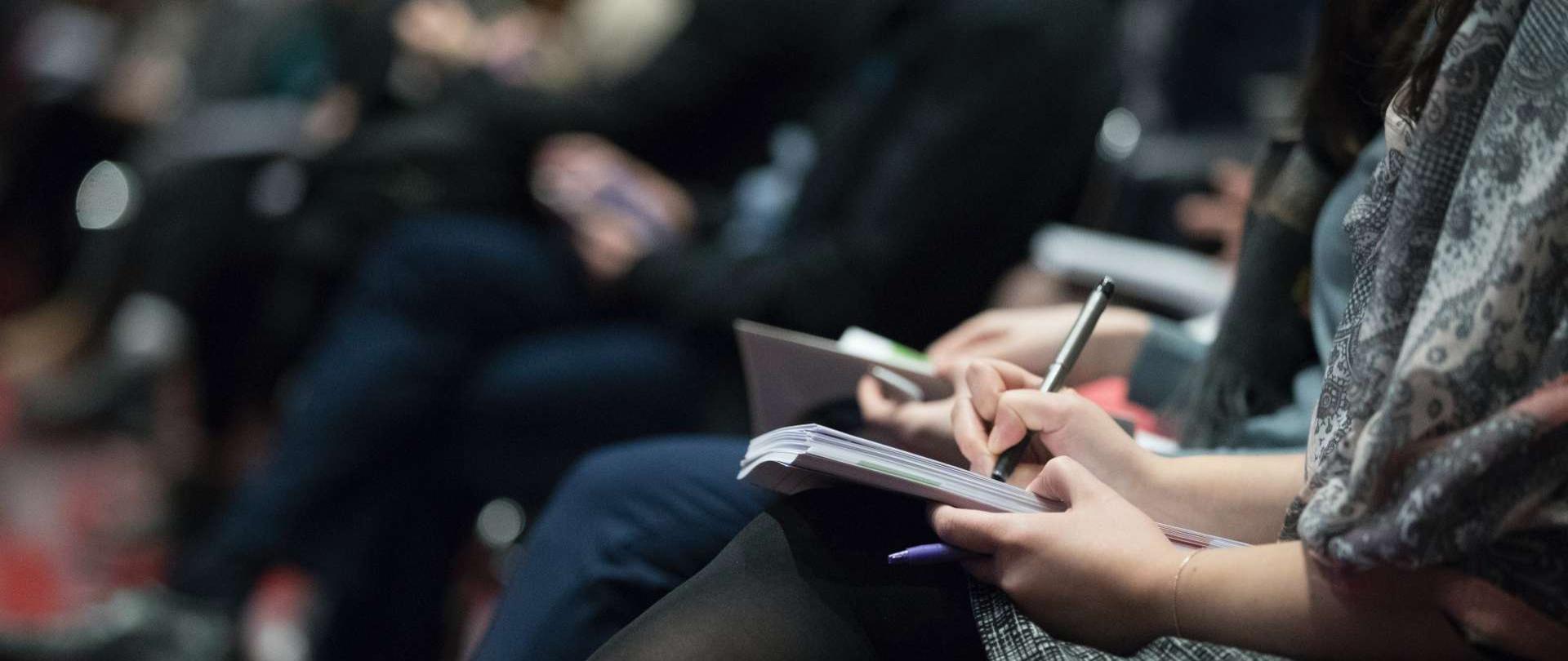 fotografia przedstawia sylwetki siedzących osób, które trzymają w dłoniach notesy i długopisy