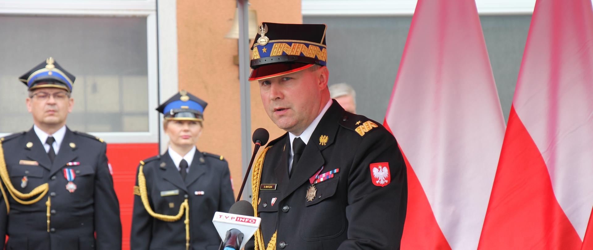 strażak w rogatywce i w mundurze ze sznurem przemawia stojąc przy mównicy pod czerwonym namiotem