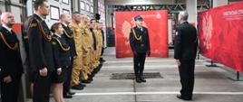 Strażacy stoją ustawieni w dwuszeregu w mundurach czarnch i zółtych, dwóch z nich stoi na środku przed nimi ustawieni twarzami do siebie. W tle ścianka firmowa PSP.