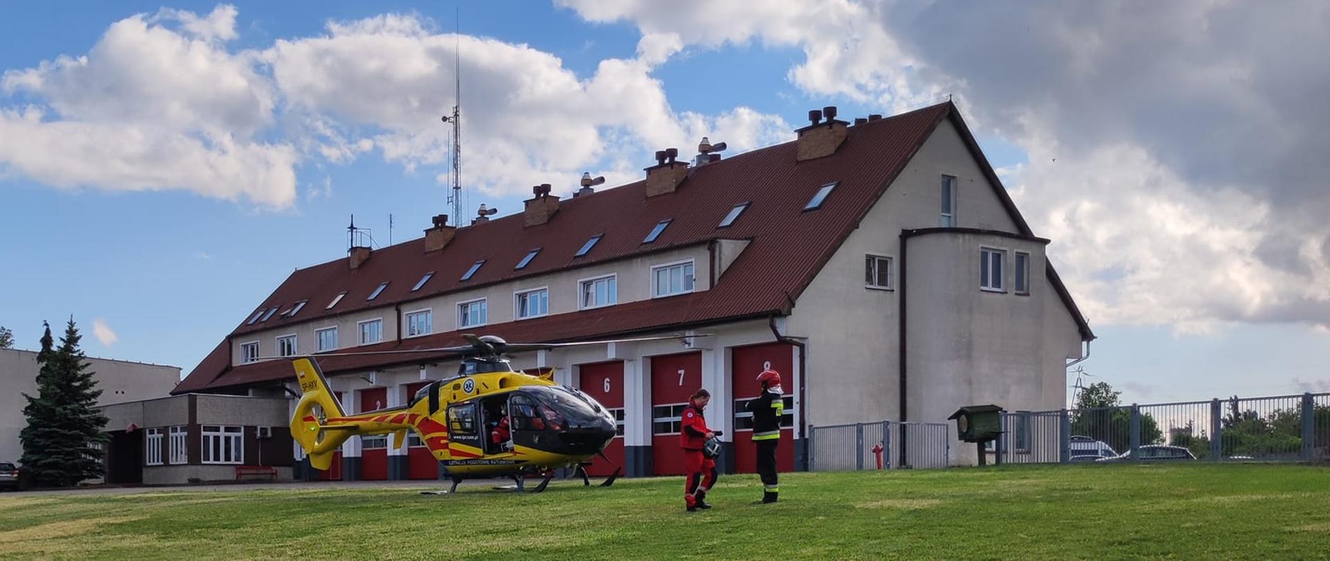 Śmigłowiec LPR wylądował na trawniku przy komendzie. Obok stoi strażak i ratownik medyczny.
