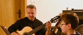Zdjęcie przedstawia od lewej strony mężczyznę trzymającego gitarę oraz ucznia obserwującego zapis nutowy na pulpicie do nut na tle drzwi do sali i beżowej ściany.