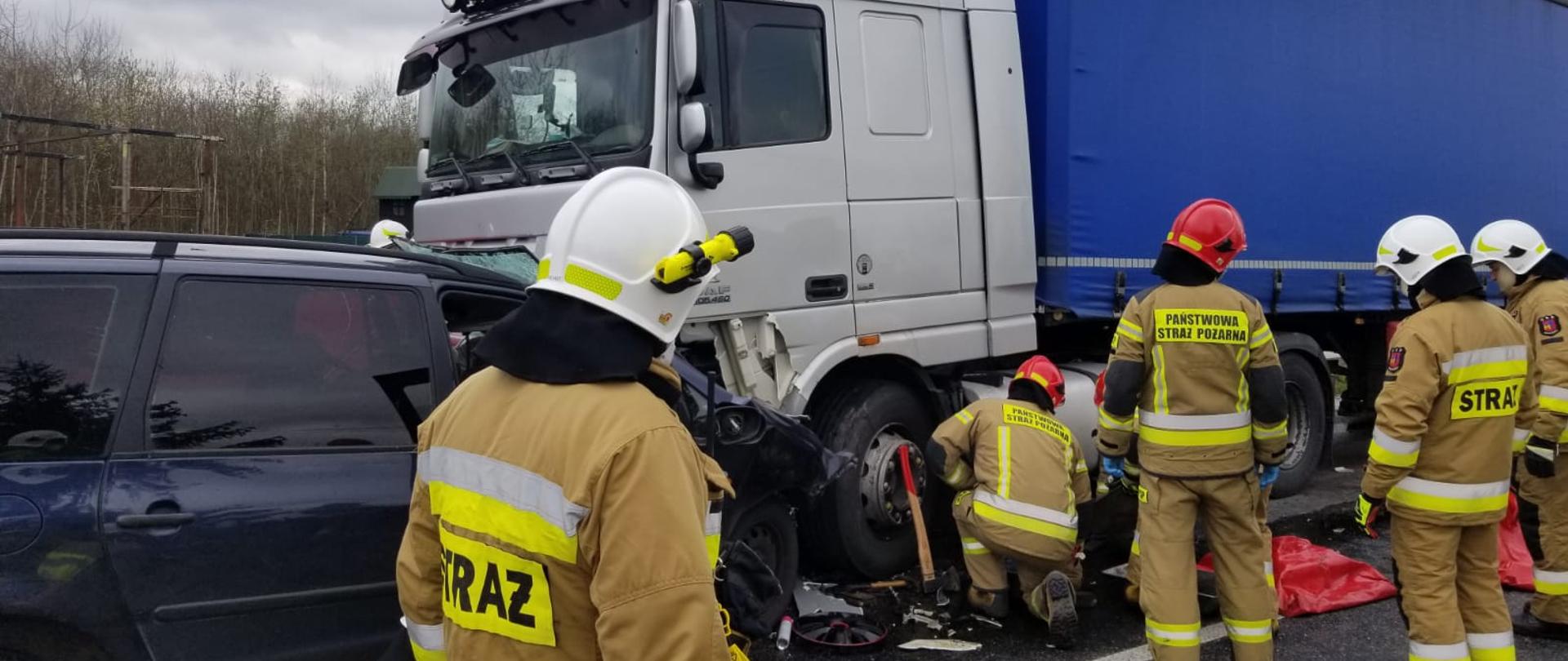 Zdjęcie przedstawia zderzenie czołowe dwóch pojazdów osobowego z ciężarowym. Przy pojazdach znajdują się strażacy ubrani w żółte ubrania specjalne. Na głowach mają czerwone oraz białe hełmy. Strażacy podejmują działania związane z ograniczeniem wycieku paliwa z uszkodzonego zbiornika poprzez podstawienie pod zbiornik plastikowych pojemników. 