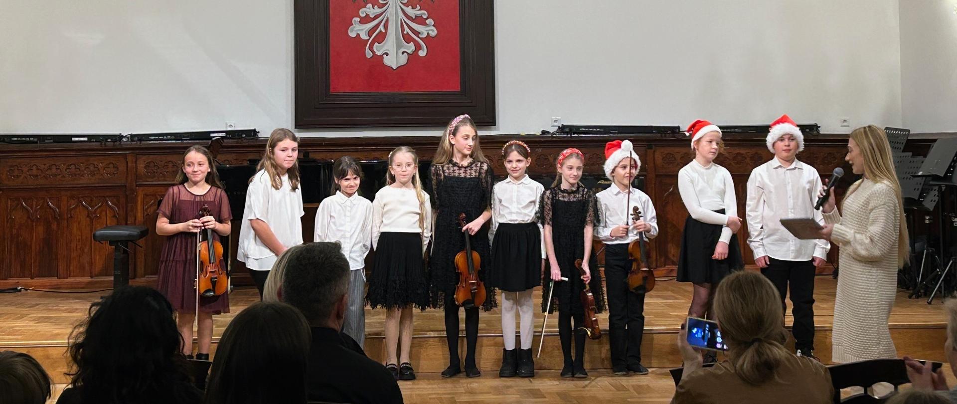Grupa dzieci stojących na scenie w Sali Królewskiej, część z nich trzyma skrzypce, po prawej stronie nauczycielka z mikrofonem mówi do uczniów.
