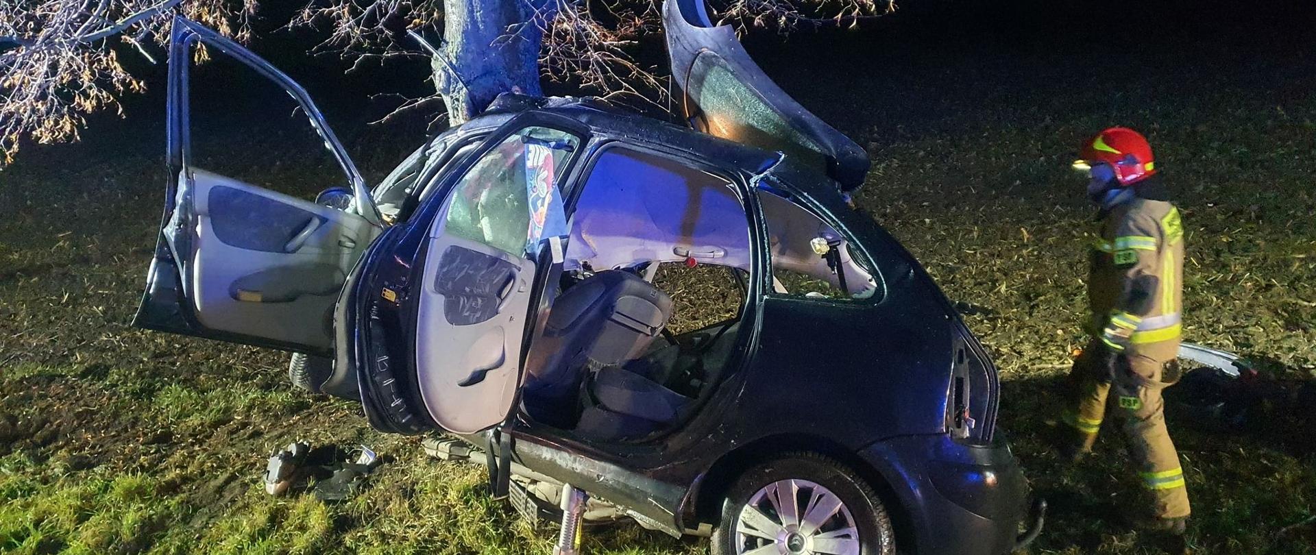 Zdeformowany samochód osobowy, który uderzył w drzewo. Siła uderzenia była tak duża, że przód pojazdu został bardzo mocno uszkodzony. Obok pojazdu stoi strażak. Samochód marki Citroen znajduje się w rowie poza jezdnią
