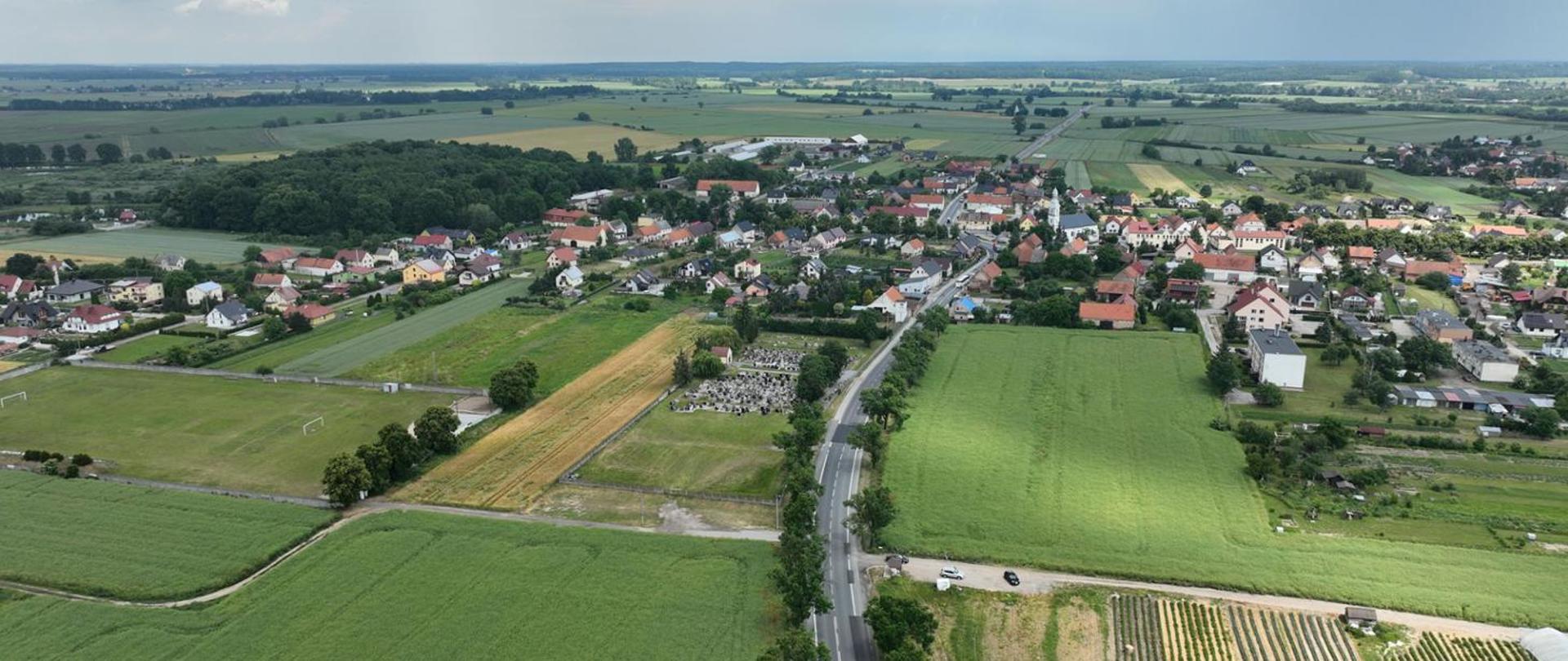 Panorama małej miejscowości otoczonej polami uprawnymi, widok z lotu ptaka. W centrum widać zabudowania mieszkalne, kościół z wieżą, a wzdłuż drogi biegnącej przez pola znajduje się cmentarz. W tle rozciągają się zielone pola i lasy pod jasnym, częściowo zachmurzonym niebem.