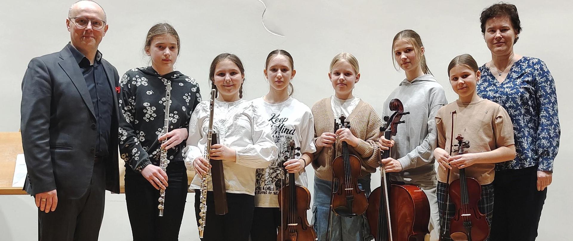 Zdjęcie przedstawia stojących zespół kameralny z prowadzącym warsztaty dr Janem i naucz. Dianą Gabor Romanowskim, uczniowie trzymają instrumenty - flety, skrzypce i wiolonczelę, za nimi profil Chopina na ścianie