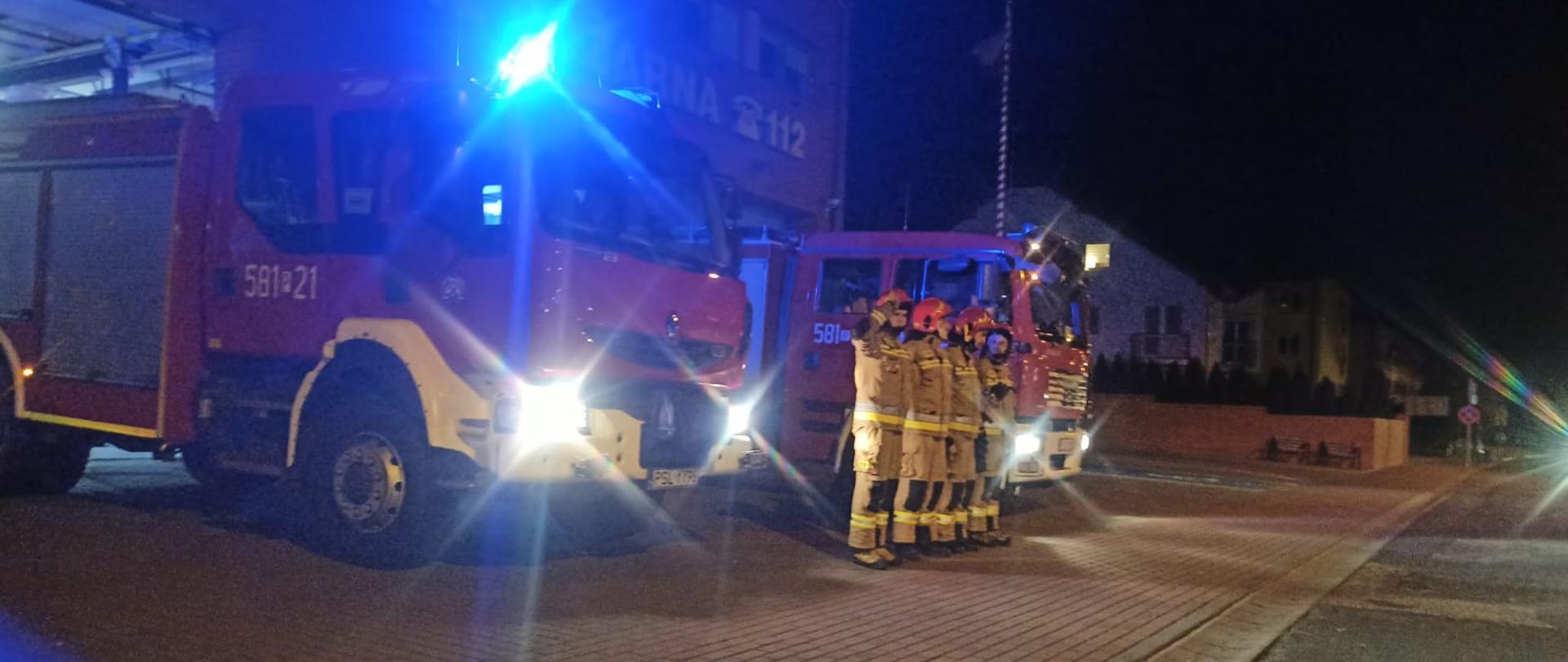 Strażacy ubrani w ubrania specjalne koloru piaskowego stoją pomiędzy dwoma samochodami pożarniczymi z włączonymi światłami błyskowymi oraz drogowymi na tle budynku Komendy PSP w Słupcy w oddali maszt.