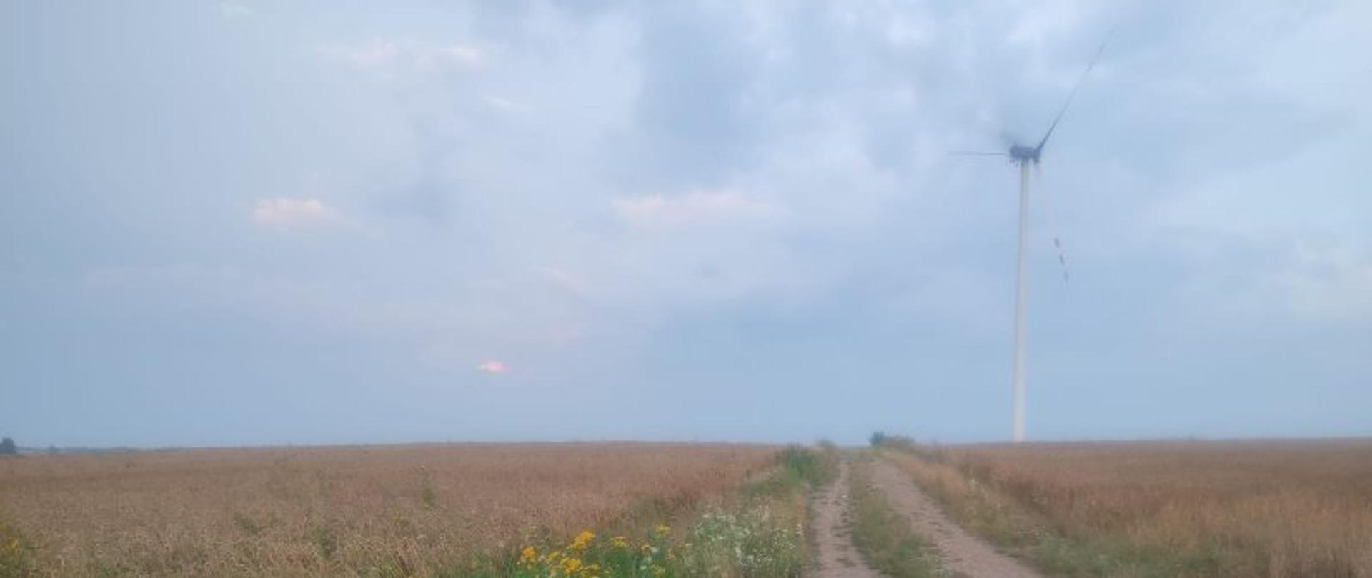 Na pierwszym planie widoczna droga polna oraz pola uprawne. Na drugim planie widoczna turbina wiatrowa. Z turbiny wiatrowej unosi się dym.