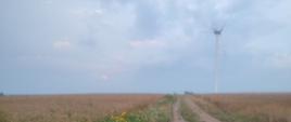 Na pierwszym planie widoczna droga polna oraz pola uprawne. Na drugim planie widoczna turbina wiatrowa. Z turbiny wiatrowej unosi się dym.