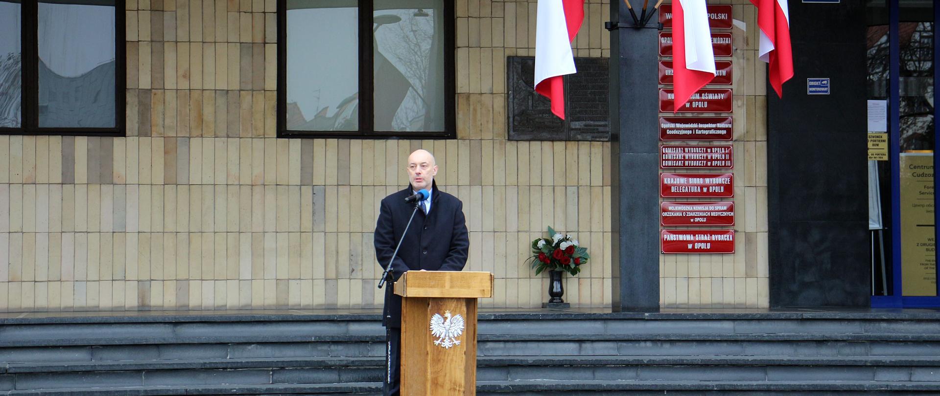 Na zdjęciu w centralnej jego części za drewnianą mównicą, stoi wojewoda opolski który przemawia do zgromadzonych przed Urzędem. W tle budynek urzędu. Po prawej stronie kadru na jednym ze słupów urzędu widać trzy biało-czerwone flagi RP. 
