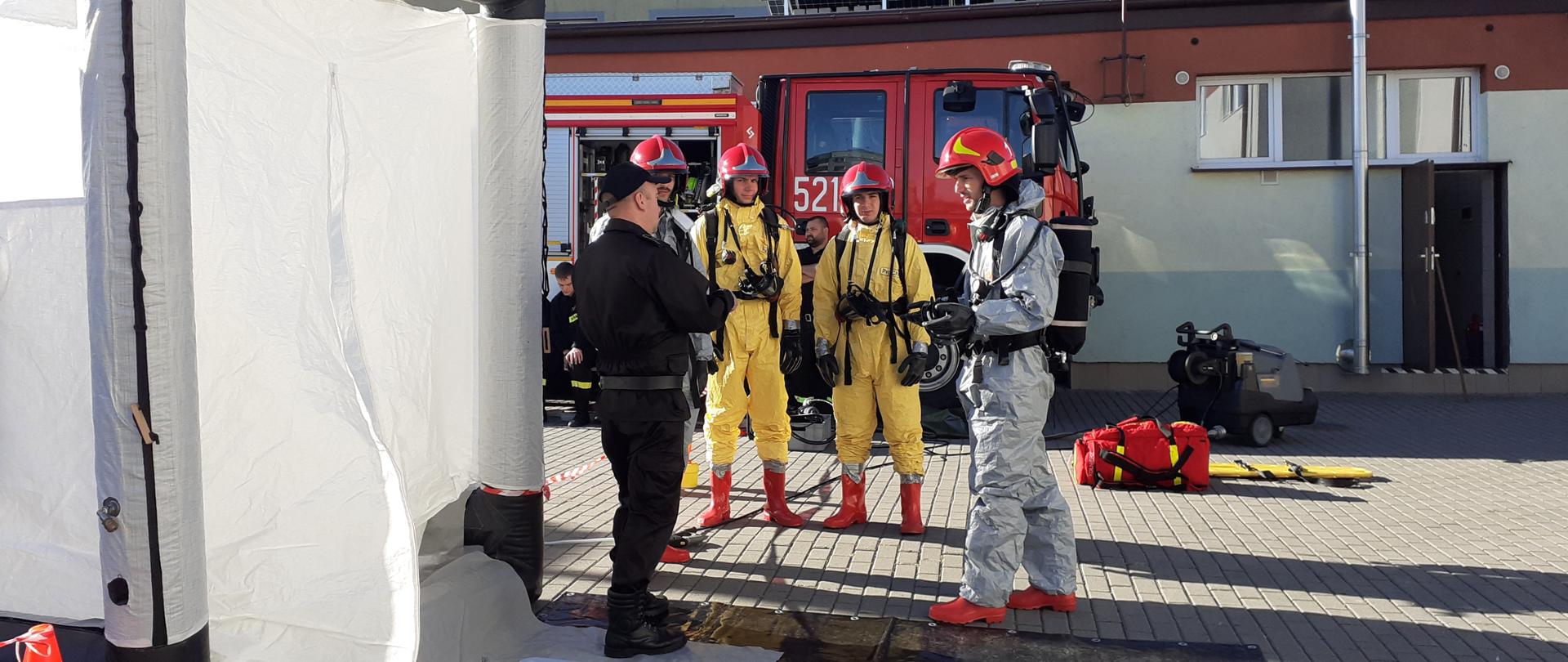 Strażak ubrany w czarny mundur przekazującego założenia do realizacji zadań z zakresu dekontaminacji dla czterech strażaków stojących przed nim obok jest rozłożony sprzęt.