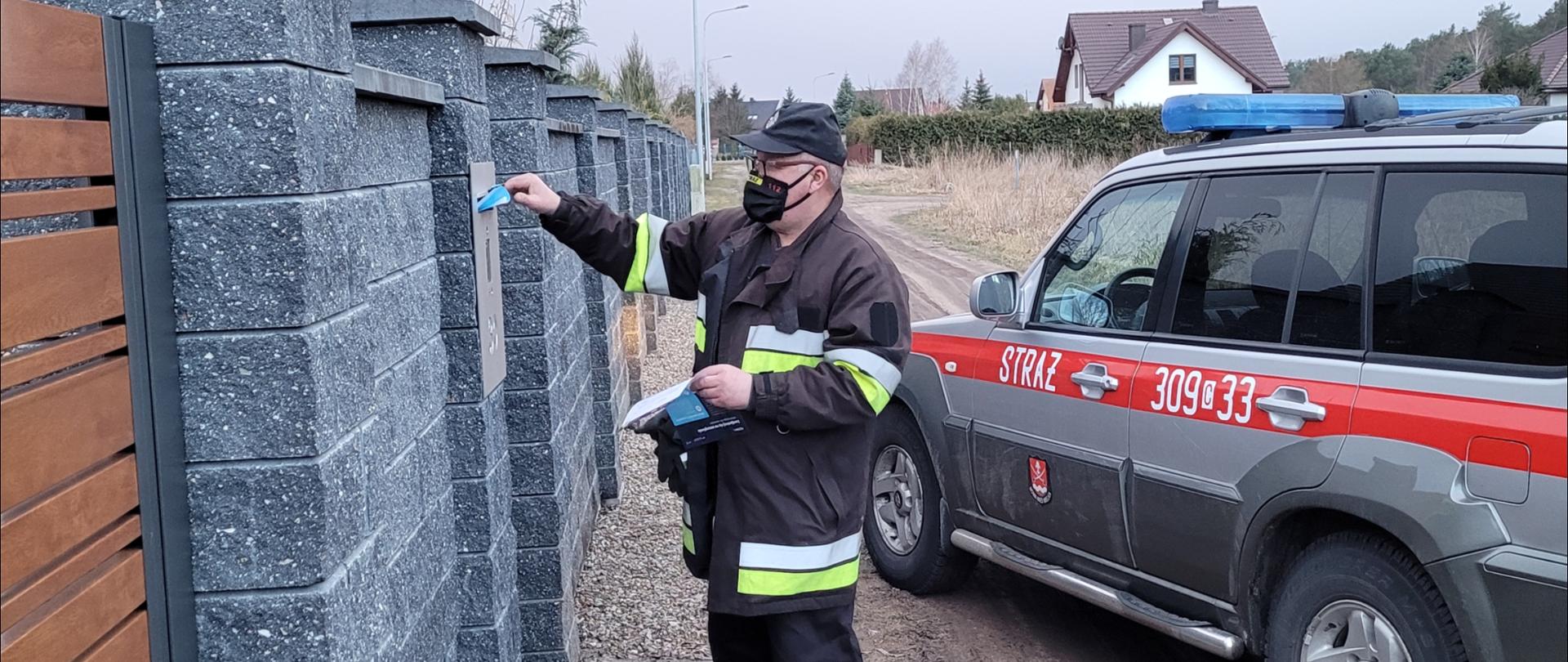 W centralnej części zdjęcia strażak stojący przy płocie posesji w ubraniu specjalnym, na głowie czarna czapka z daszkiem, na twarzy maseczka. Strażak wrzuca ulotkę do skrzynki pocztowej, która jest na płocie. W prawej części zdjęcia stoi samochód pożarniczy osobowy koloru siwego z czerwonym paskiem należący do OSP Nowa Wieś Wielka